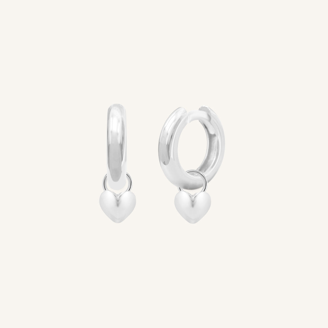 A pair of silver-colored hoop earrings with a dangling heart charm.