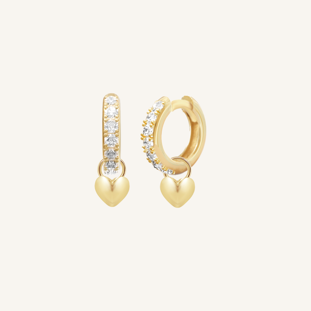 A pair of gold hoop earrings with cubic zirconias with heart charms attached to them, displayed against a white background.