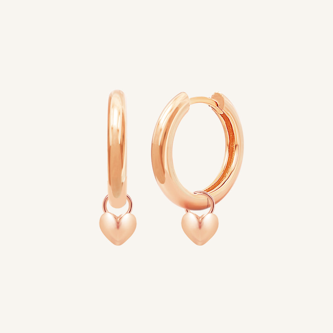 A pair of rose-colored hoop earrings with a dangling heart charm.