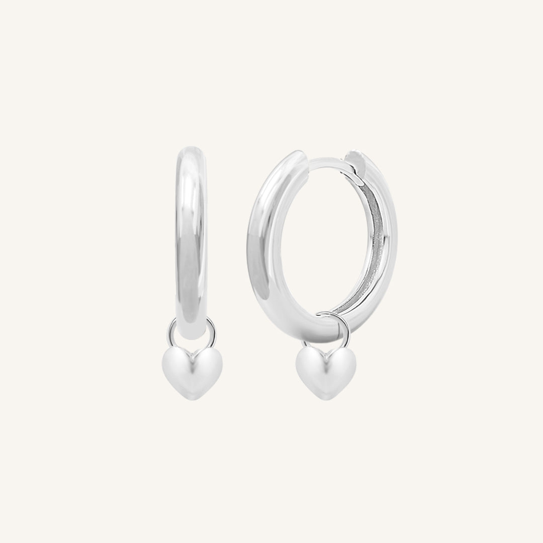 A pair of silver-colored hoop earrings with a dangling heart charm.