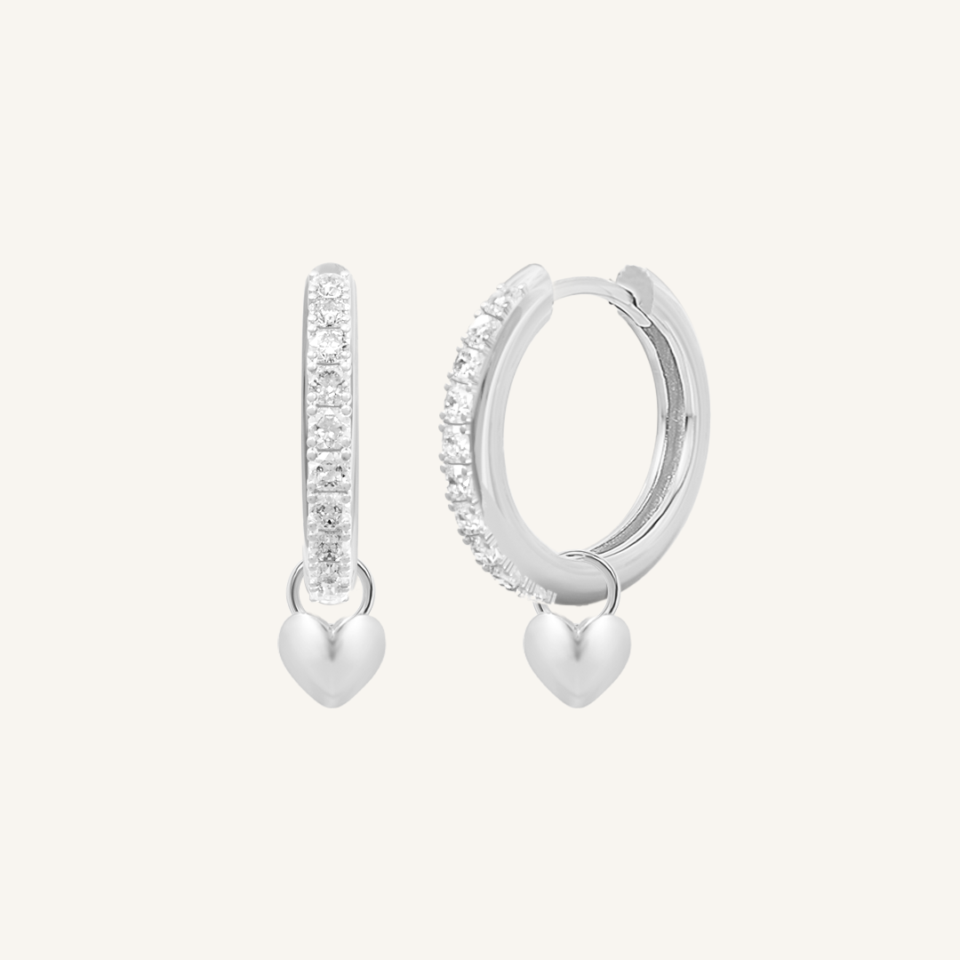 A pair of silver hoop earrings with cubic zirconias with heart charms attached to them, displayed against a white background.