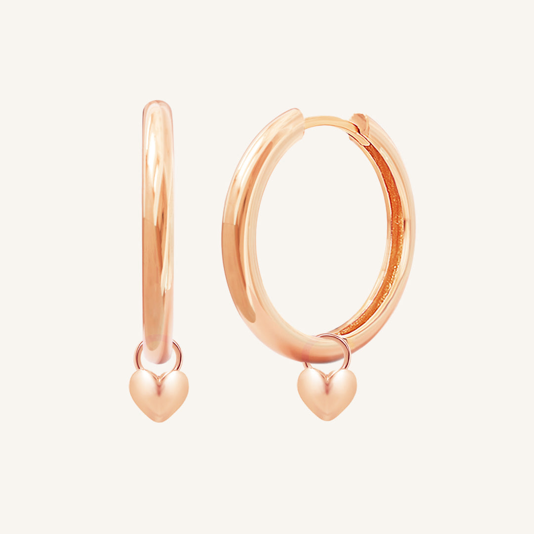 A pair of rose-colored hoop earrings with a dangling heart charm.