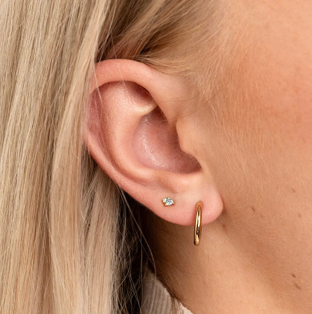 A gold pair of stud earrings featuring a white cubic zirconia, representing April, worn in ears.