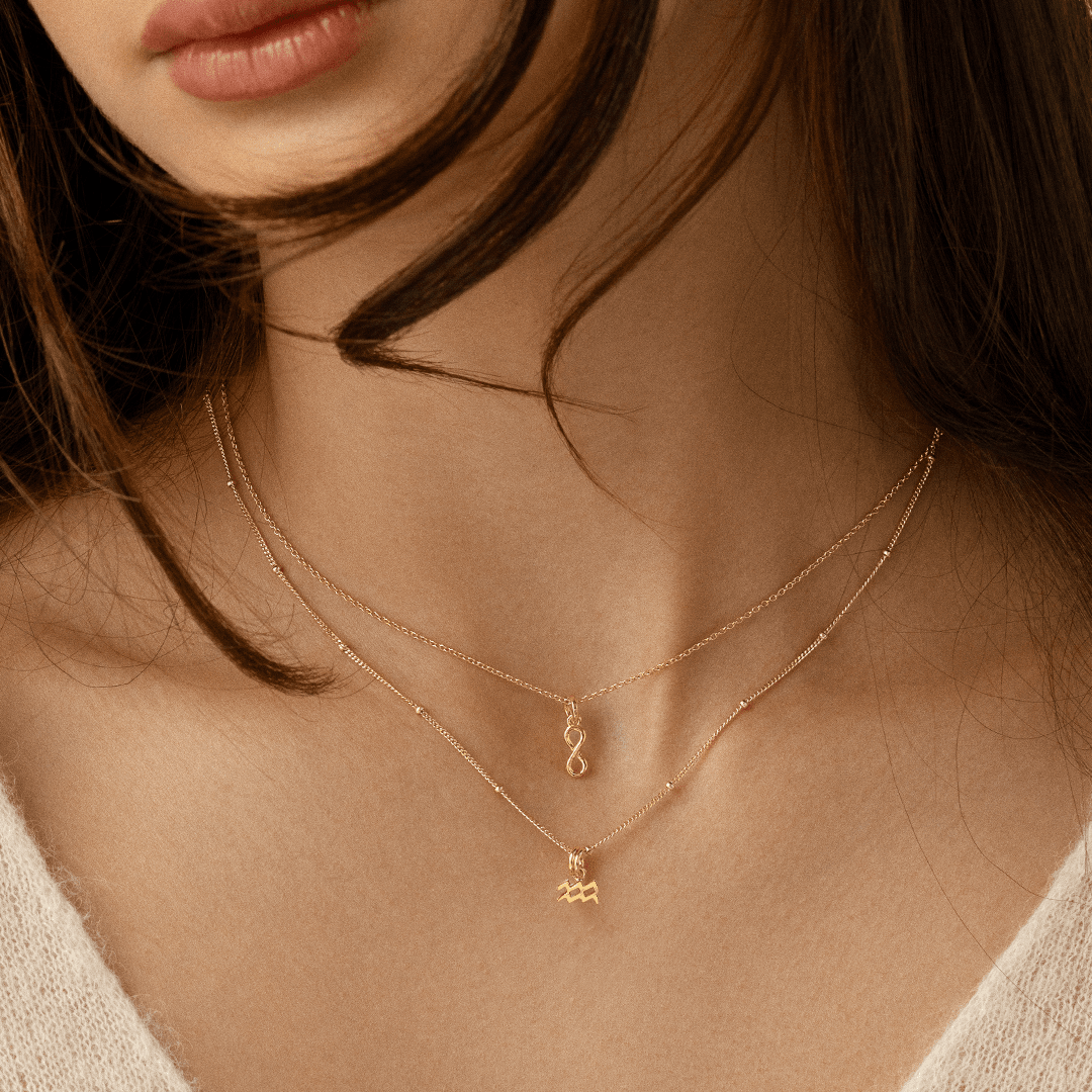 A gold-coloured Petite Zodiac Charm representing the Aquarius sign on a chain around the neck