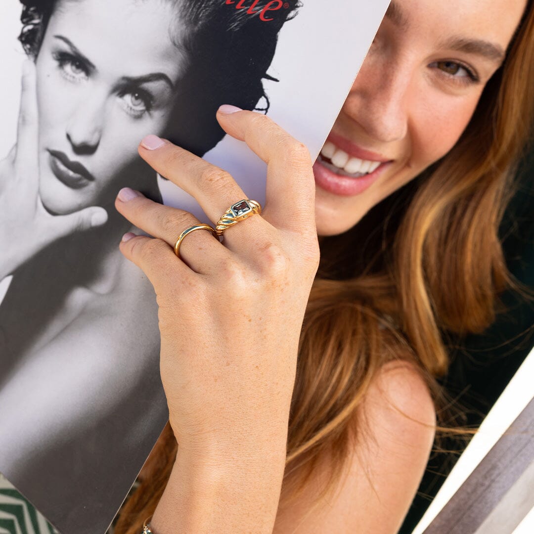 Woman wearing a gold ring with a gemstone, with a vintage-style poster in the background.