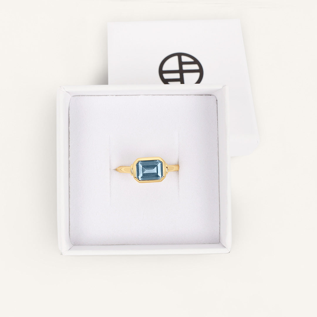 Gold ring with a blue gemstone in a white box with a brand logo.