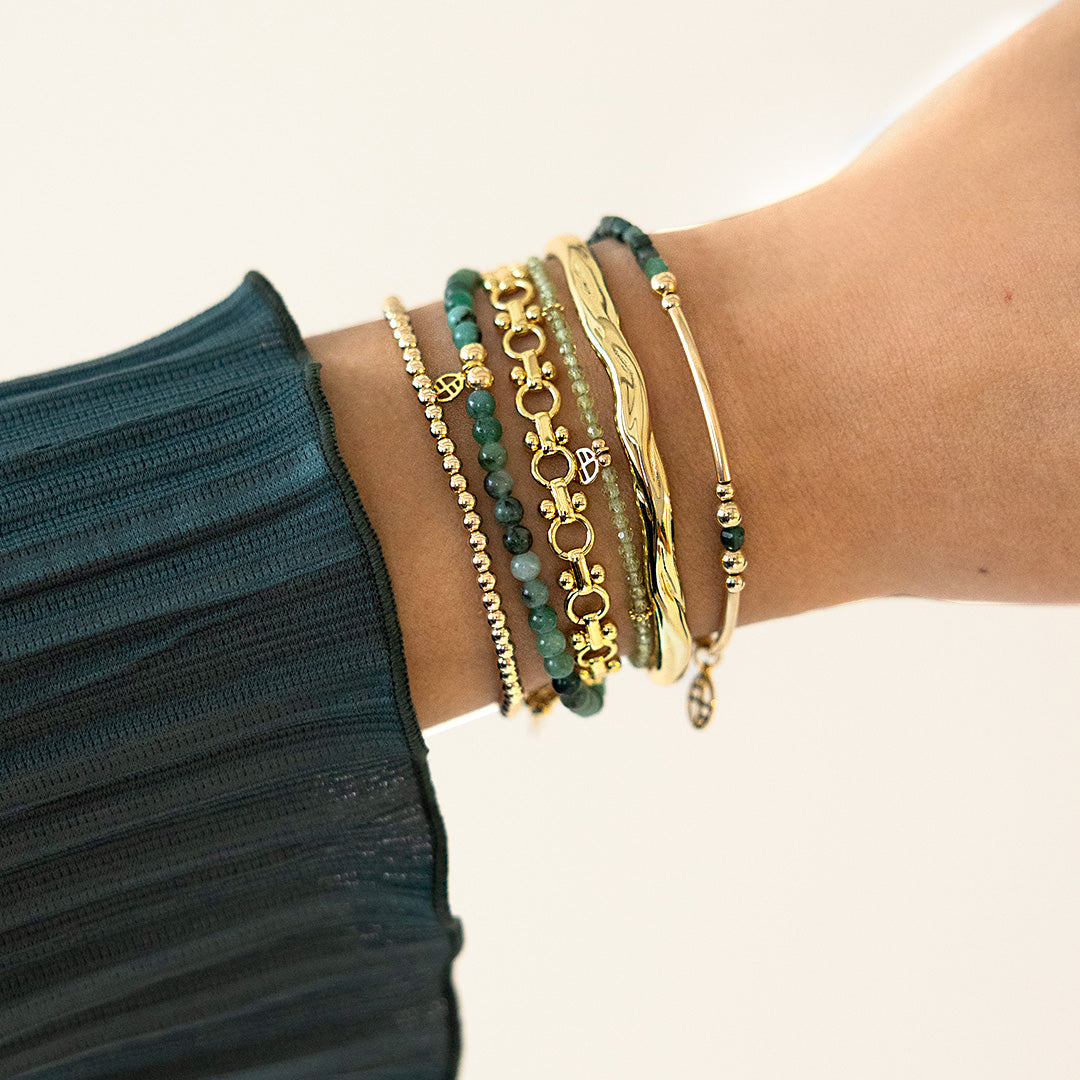 Close-up of a wrist wearing multiple gold and green beaded bracelets on a neutral background