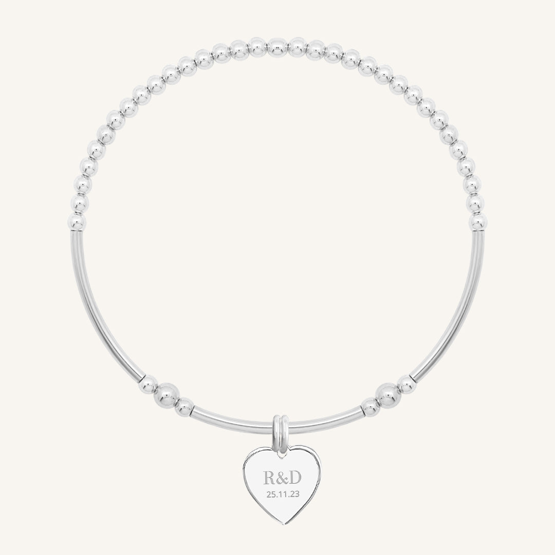 A silver bracelet with a beaded design and a heart-shaped charm, engraved with initials and a date