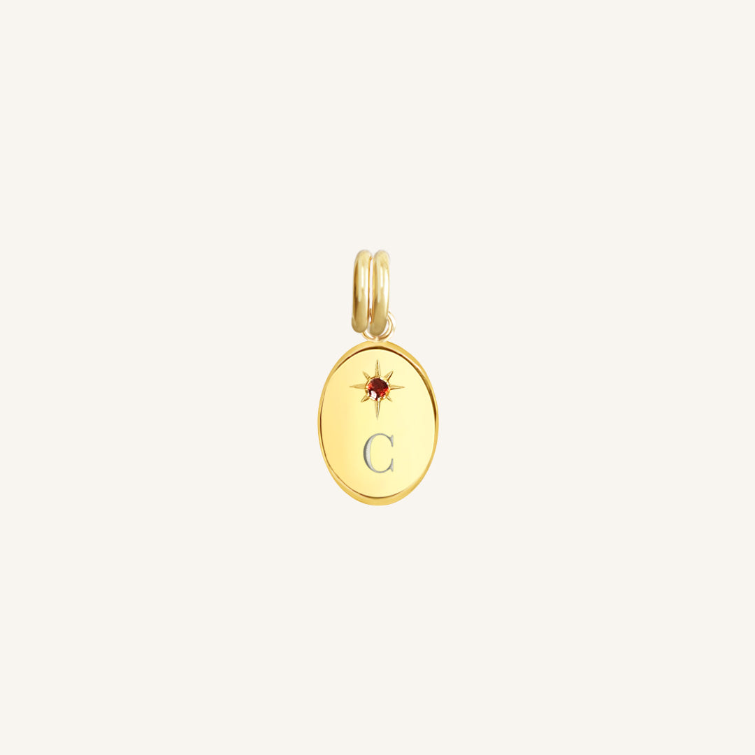 Gold pendant with letter 'C' and red stone on a light beige background