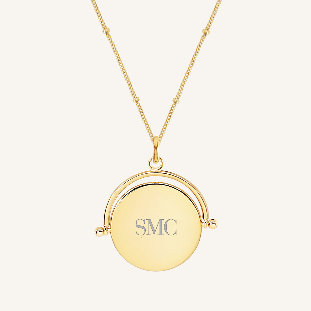 A gold-coloured pendant with the initials engraved on it, featuring a spinning mechanism, on gold chain
