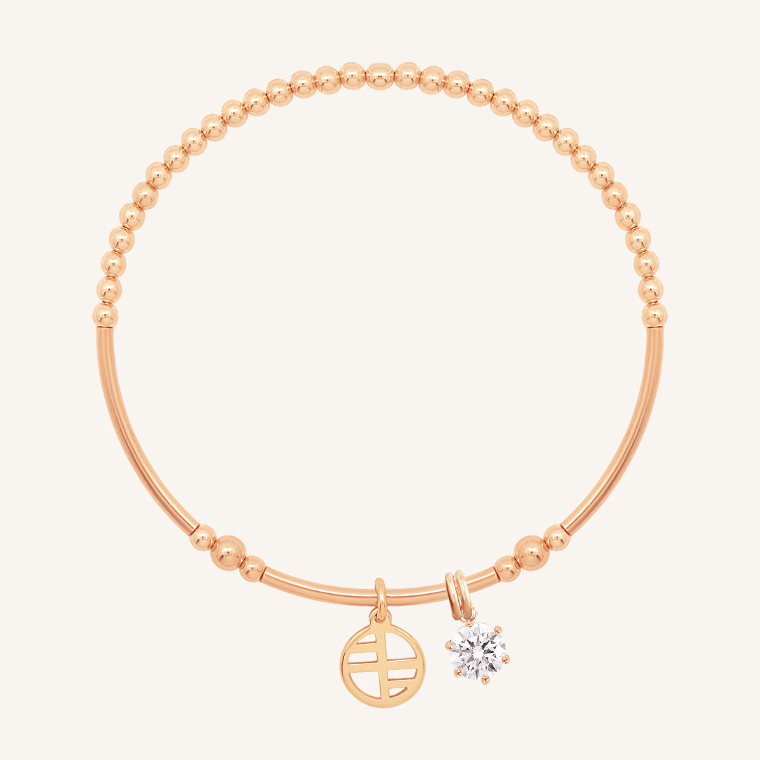 A rose-tone charm bracelet with a beaded design and two charms, one circular and one in the shape of a heart with a small cubic zirconia inside.