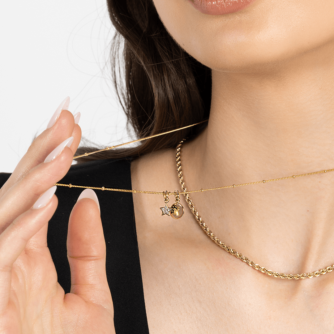 A gold coloured charm in the shape of a star with a small cubic zirconias attached to a gold chain withe an Alpha Letter Charm, around models neck