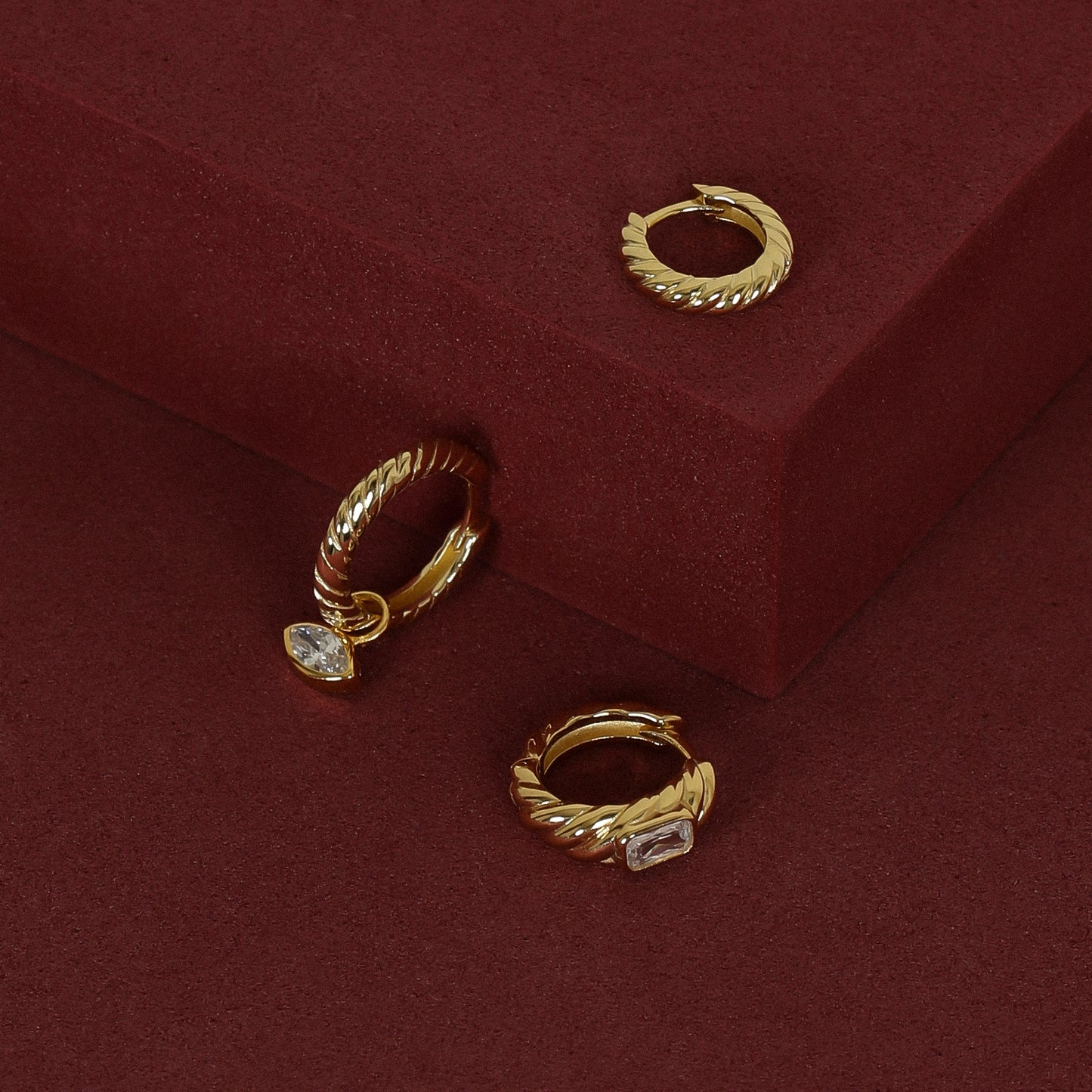 gold delicate huggies with a twist design with gold hoops inspired by the Louvre's architecture with Marquise cut white cubic zirconias with gold hoops inspired by the Louvre's architecture with Marquise cut white cubic zirconias bezel set hoop charms on red background