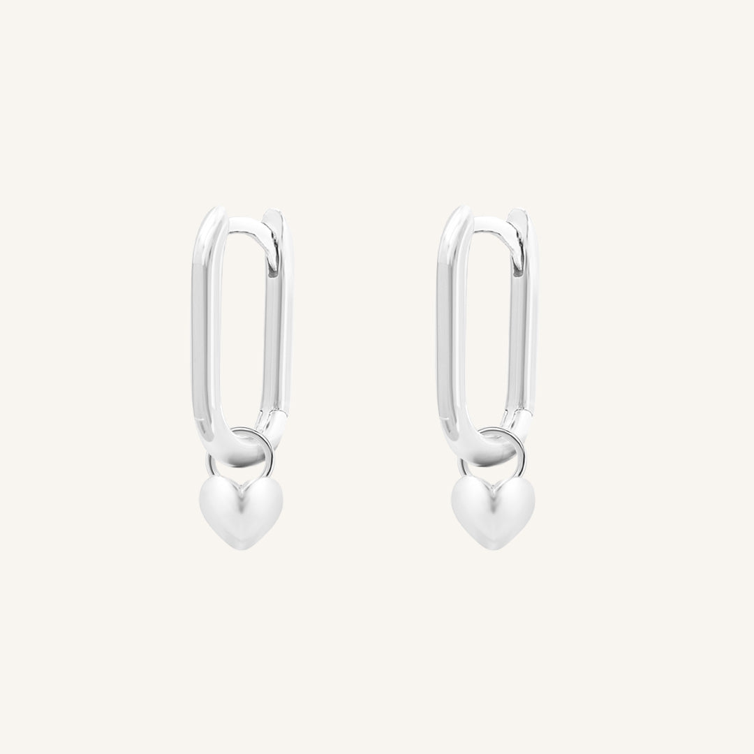 A pair of silver-tone hoop earrings with heart charms attached to them, displayed against a white background.