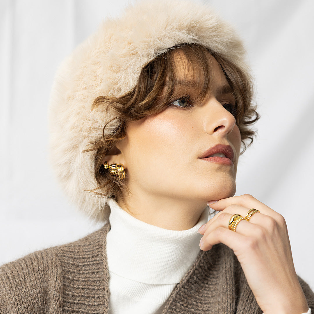 Woman wearing a fur hat, white turtleneck, and brown coat with gold jewelry against a light background