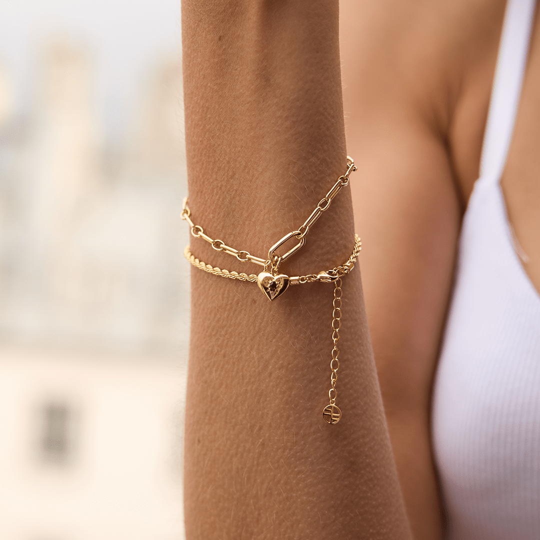 gold heart shaped charm with deep red oval cubic zirconia attached to create link bracelet on wrist with rope bracelet