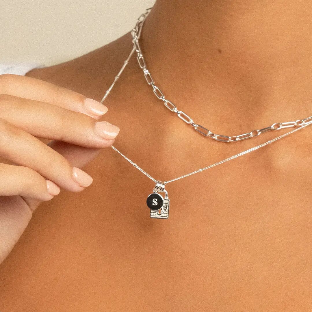 A silver-colored charm in the shape of a keylock with intricate patterns, attached to chain, on models neck