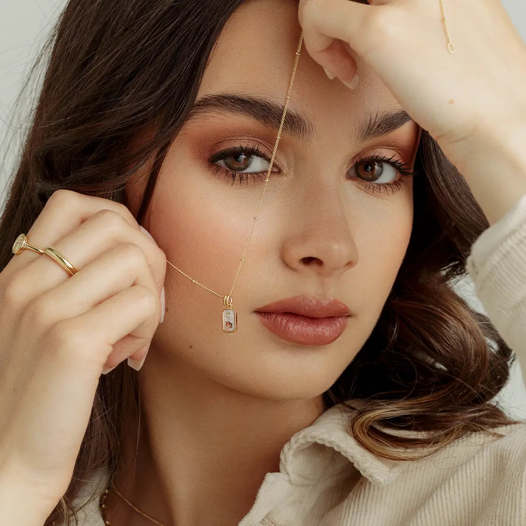 A gold iridescent zodiac charm representing the Virgo sign, with a loop for attachment on a chain, held by model
