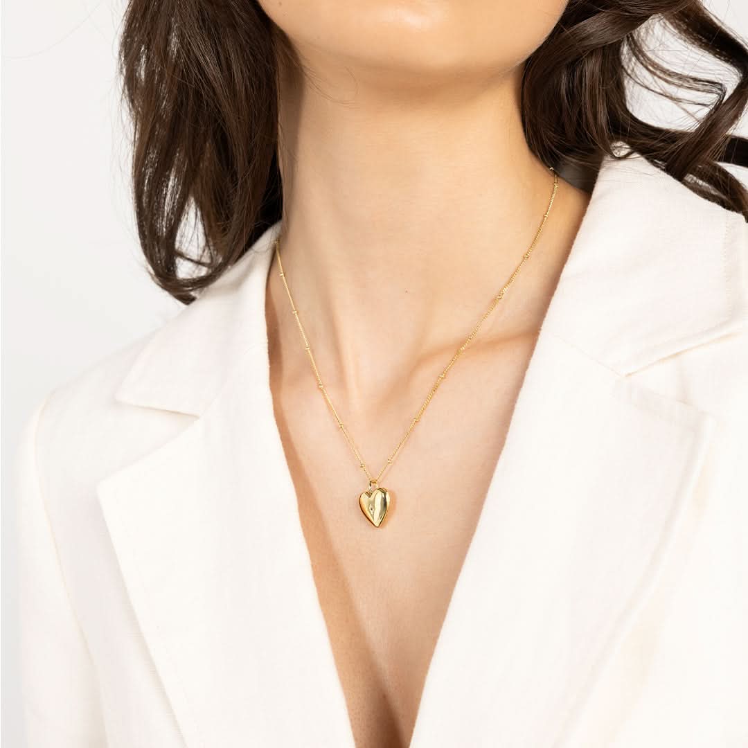 A gold heart-shaped photo locket closed on bobble chain, worn around models neck