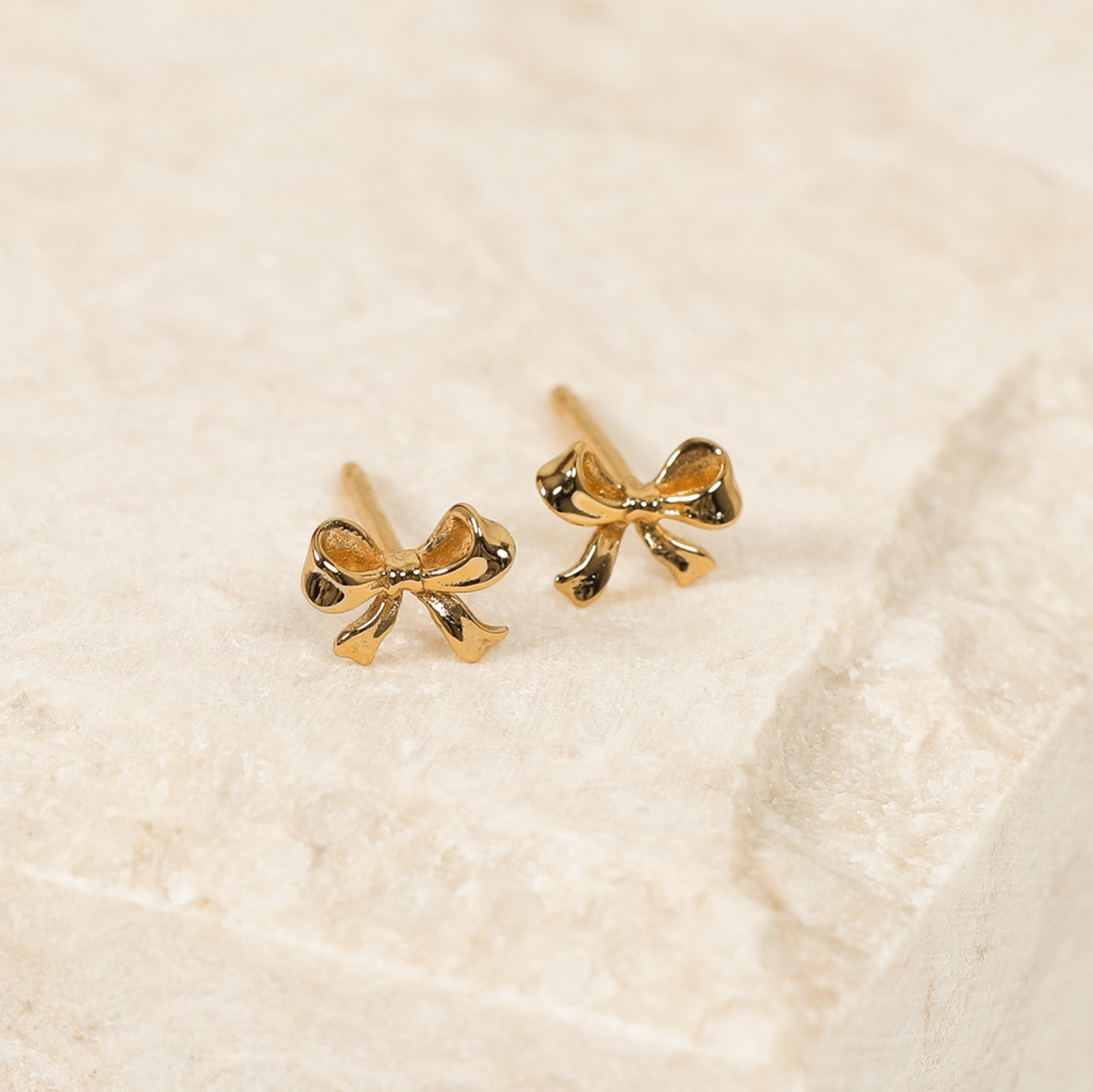 A pair of gold bow stud earrings on a beige background