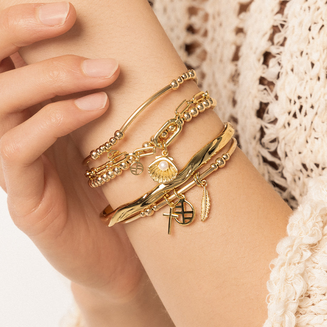 A gold-colored chain bracelet with a shell shaped charm with a pearl in the middle, worn on models wrist