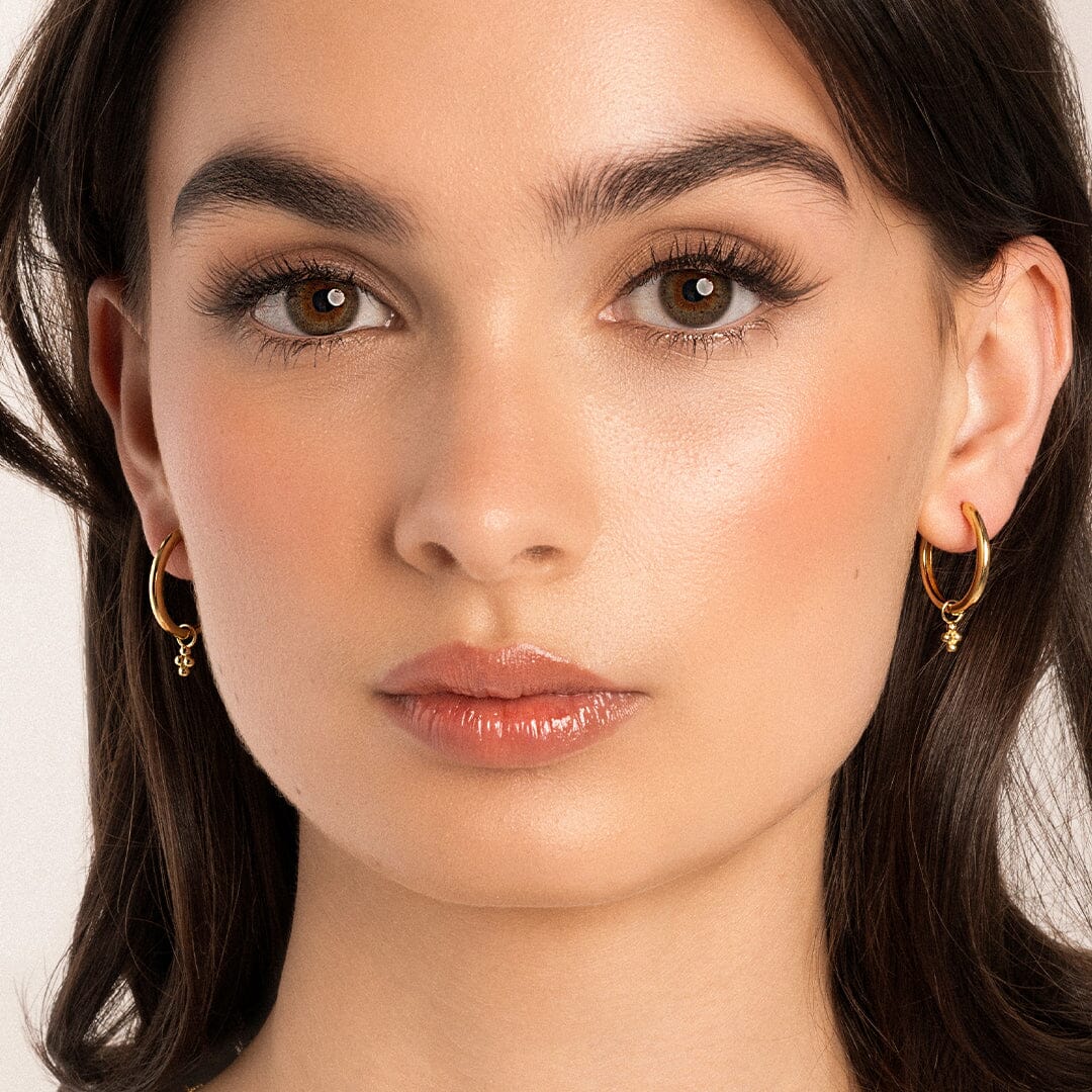 A pair of gold-tone hoop earrings with bobble details, close up in models ears