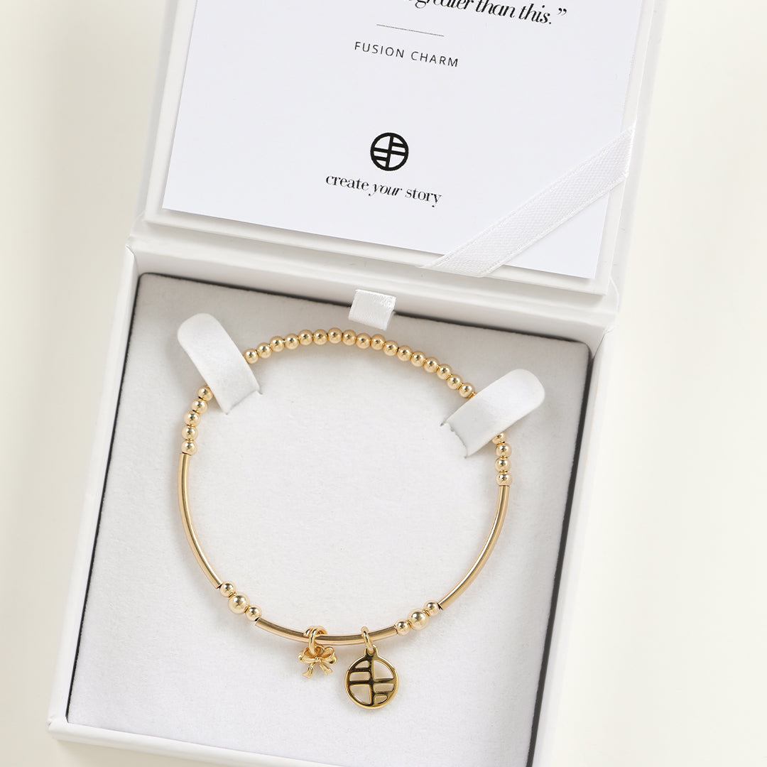 A gold-tone metal bracelet with a beaded design, featuring a charm in the shape of a bow in a white box
