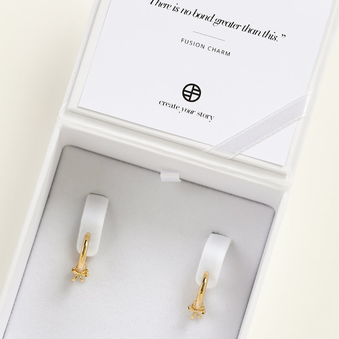 A set of two gold hoop charms with a bow design, on hoops, in box