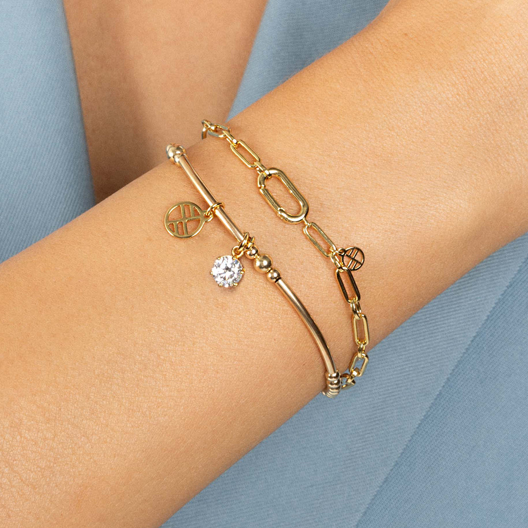 A gold-tone charm bracelet with a beaded design and two charms, one circular and one in the shape of a heart with a small cubic zirconia inside, close up on wrist.