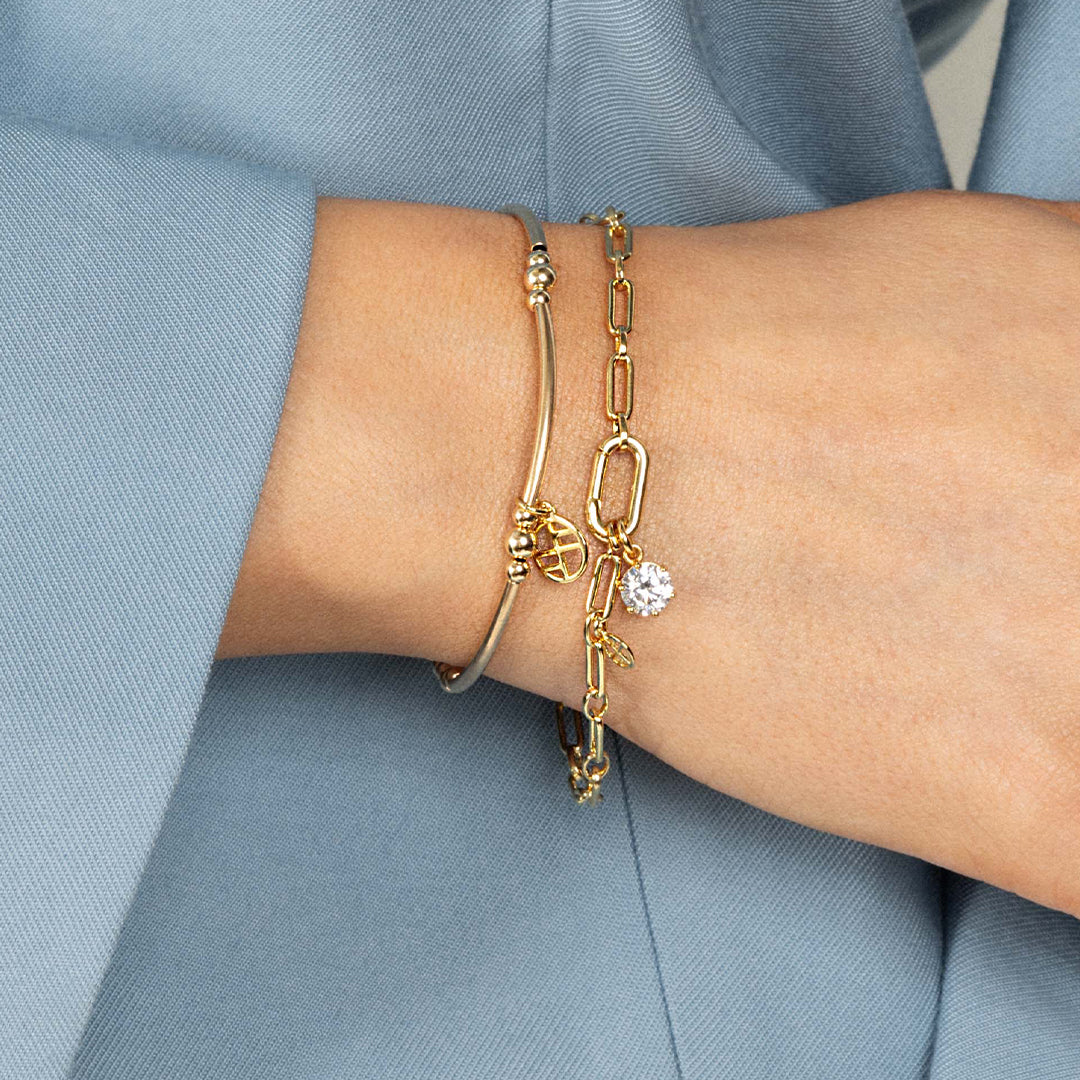 A round gold cubic zirconia charm on link bracelet, worn on wrist.