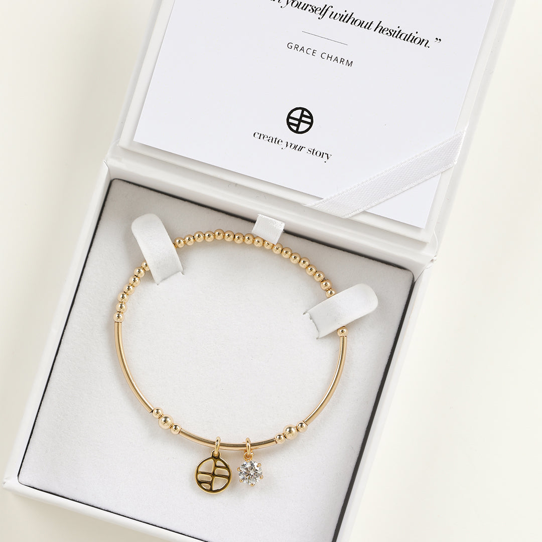 A gold-tone charm bracelet with a beaded design and two charms, one circular and one in the shape of a heart with a small cubic zirconia inside, displayed in white box.
