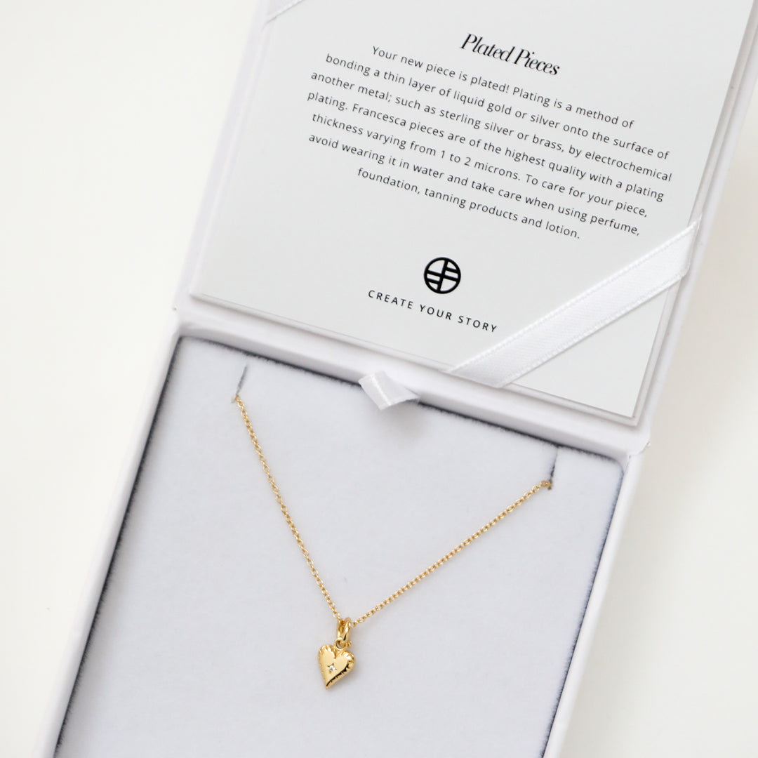 A gold-tone heart charm necklace with a fine chain, displayed in white box