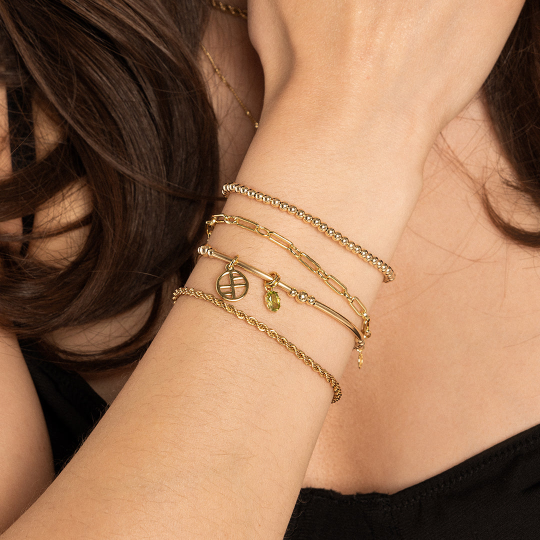 A gold charm with an olive green cubic zirconia, attached to bracelet, worn on wrist