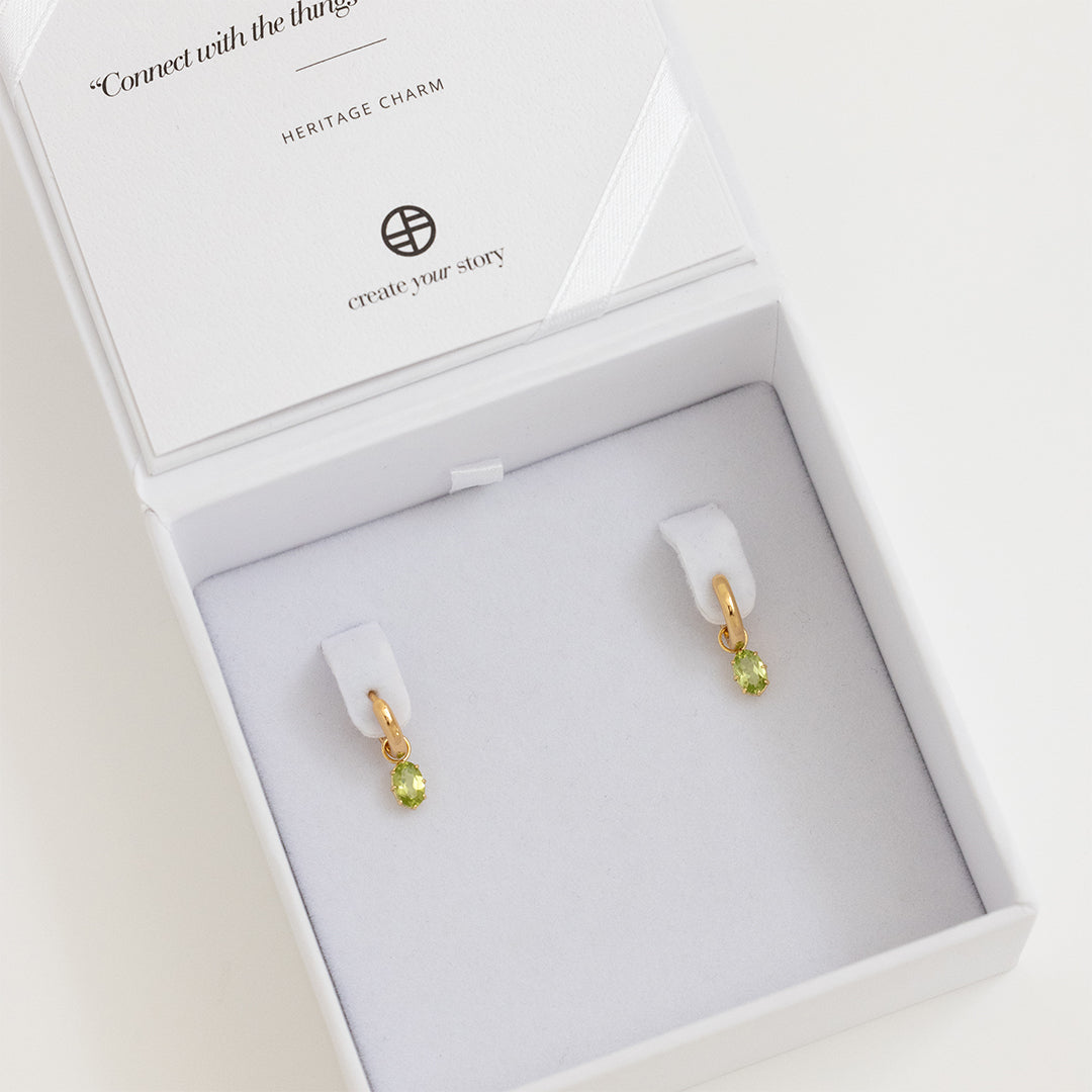 A pair of gold-tone hoop earrings with olive green charms attached to them, displayed in white box.
