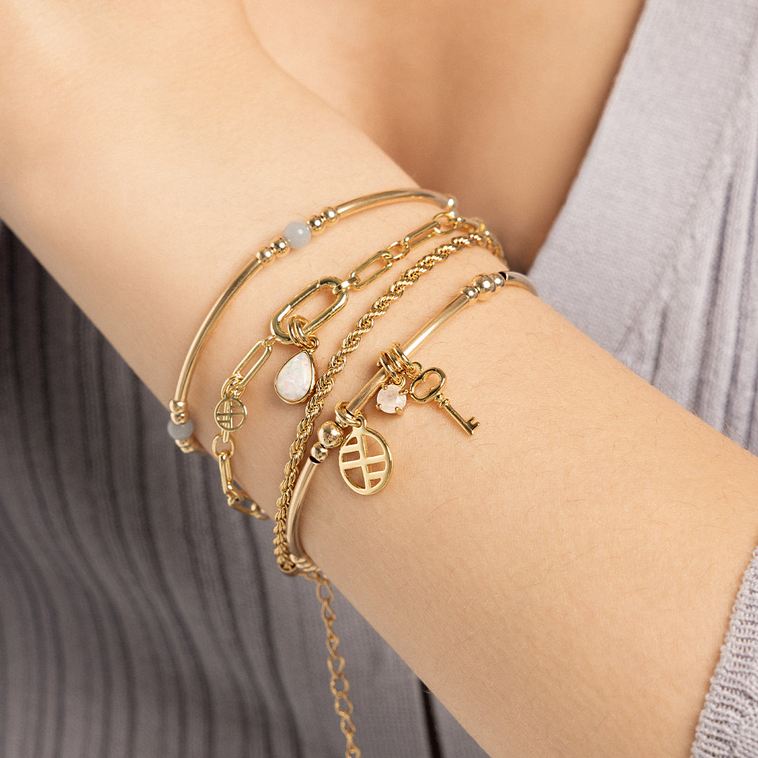 A small gold charm with a moonstone, attached to a bracelet next to a key charm, layered with bracelets on wrist