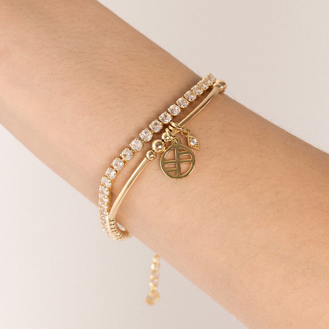 A small, diamond-shaped gold charm with a white stone in the center, attached to bracelet, on the wrist