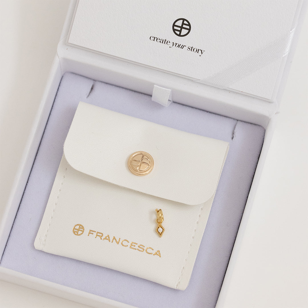 A small, diamond-shaped gold charm with a white stone in the center, in a white box.