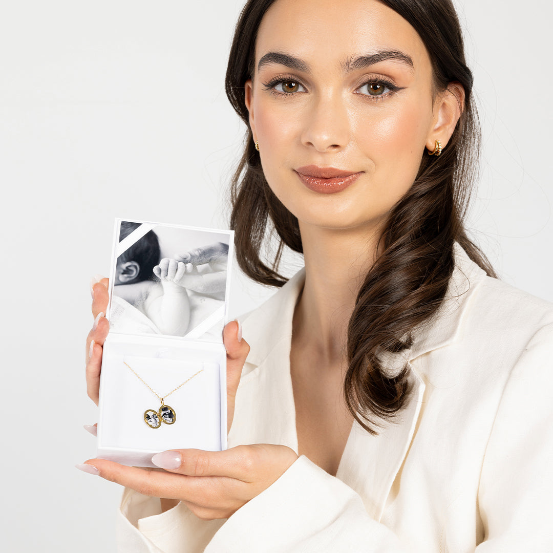 An oval-shaped photo locket with a gold metal tone, open to display two photographs, held by model