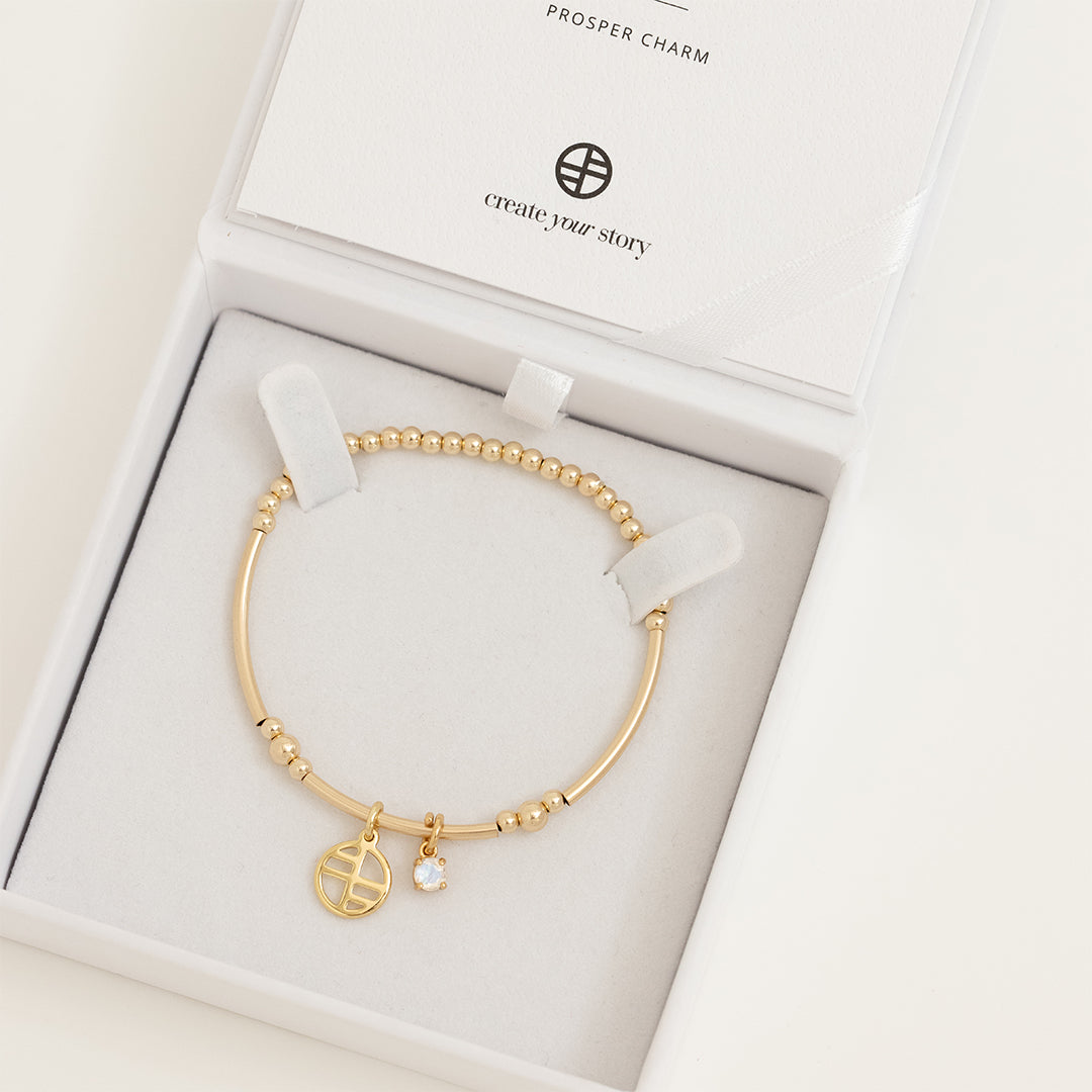 A gold-tone charm bracelet featuring a beaded design with a small circular charm and a moonstone charm, displayed in white box