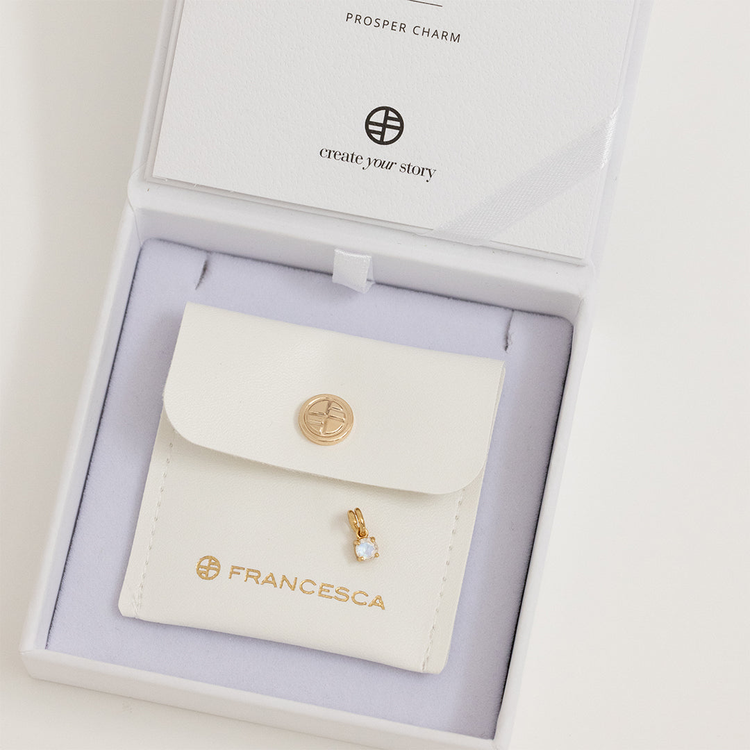 A small gold charm with a moonstone, designed to attach to a bracelet or necklace, displayed in white box
