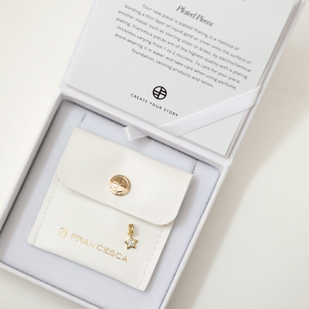 A gold coloured charm in the shape of a star with a small cubic zirconias, designed to be attached to a bracelet or chain, displayed in white box