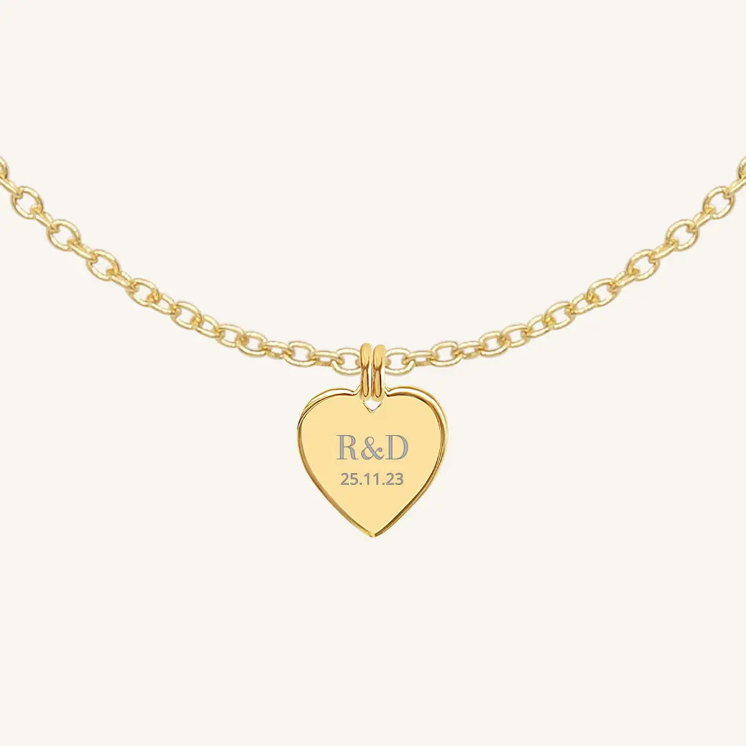 A gold heart-shaped necklace with a chain, featuring an engraved monogram initials and a date on the front.