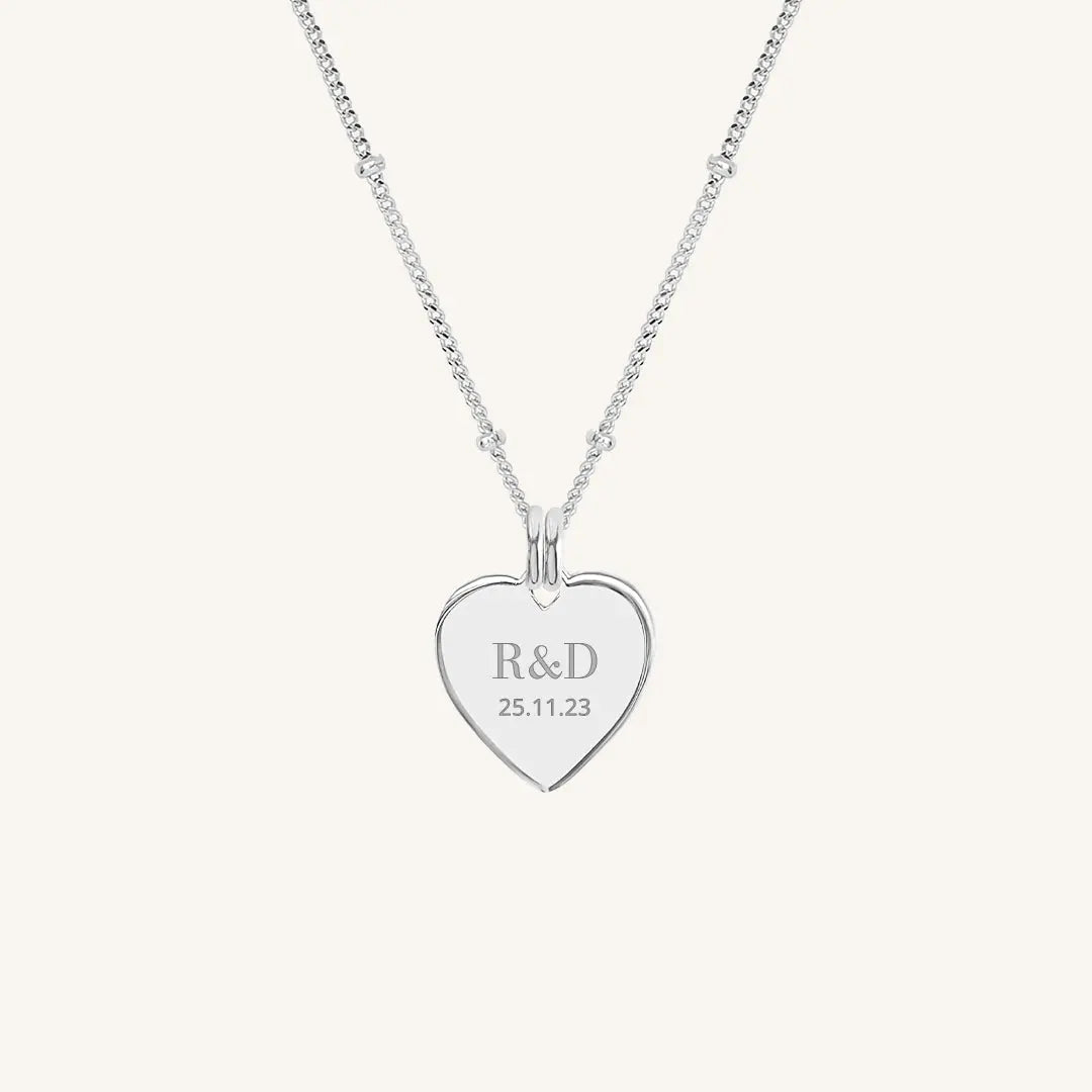A silver heart-shaped necklace with a chain, featuring an engraved monogram initials and a date on the front.