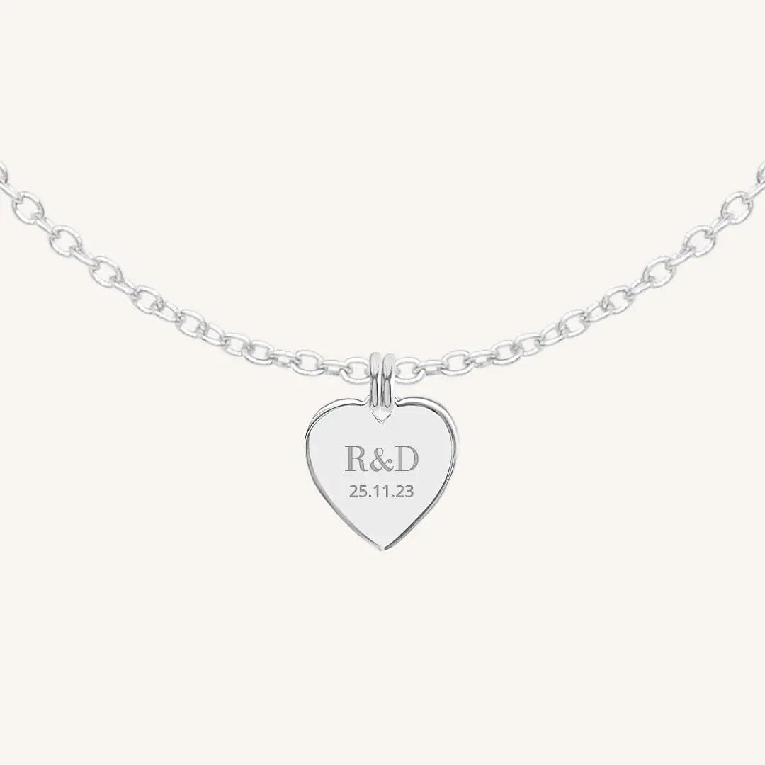 A silver heart-shaped necklace with a chain, featuring an engraved monogram initials and a date on the front.