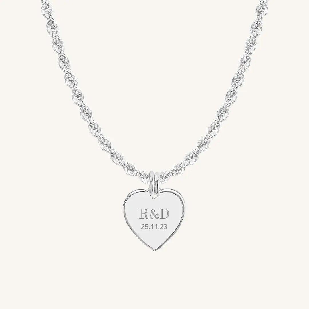 A silver heart-shaped necklace with a chain, featuring an engraved monogram initials and a date on the front.