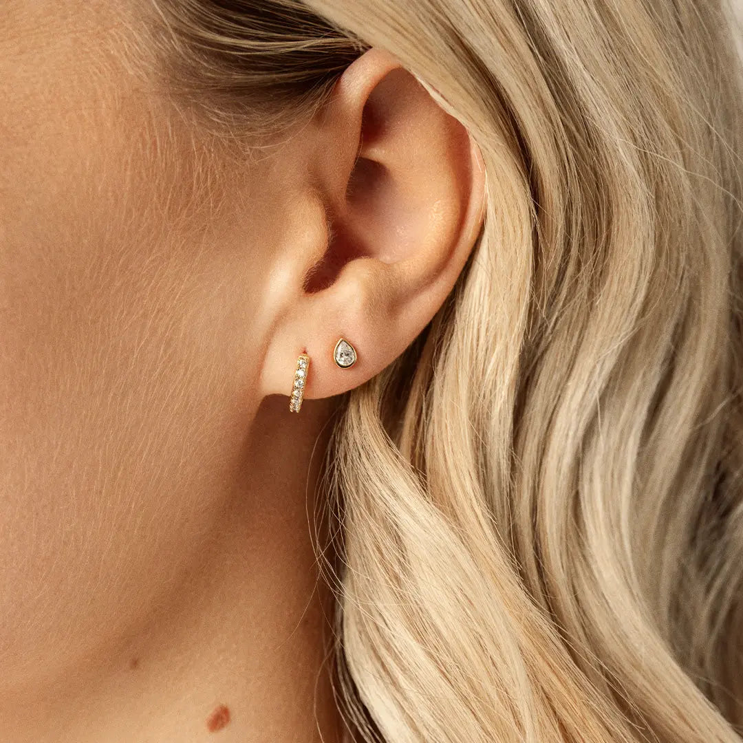 A pair of teardrop-shaped cubic zirconia stud earrings set in gold-tone settings, worn in models ear.