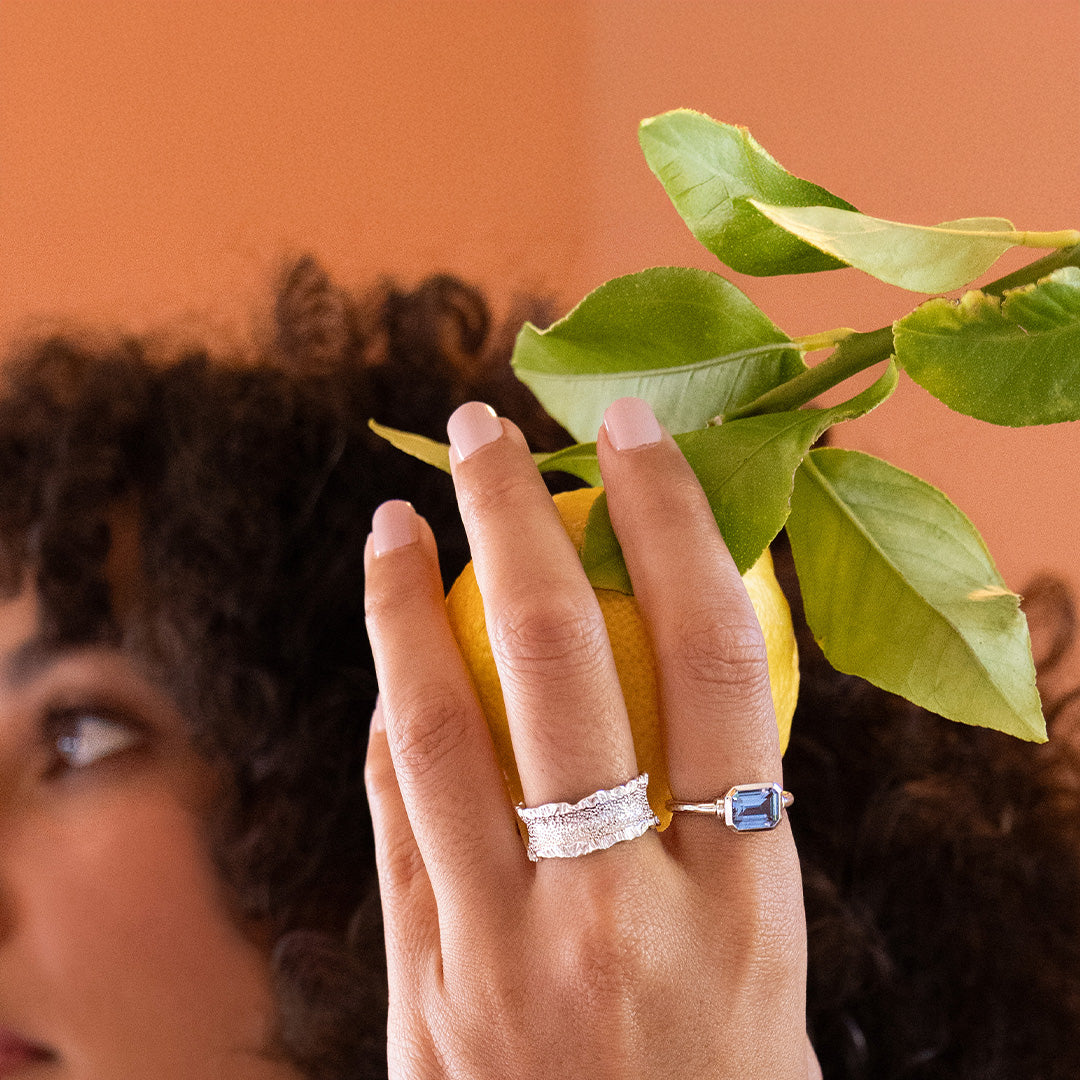Person wearing a silver ring with a blue gemstone, holding a lemon with green leaves against an orange background.