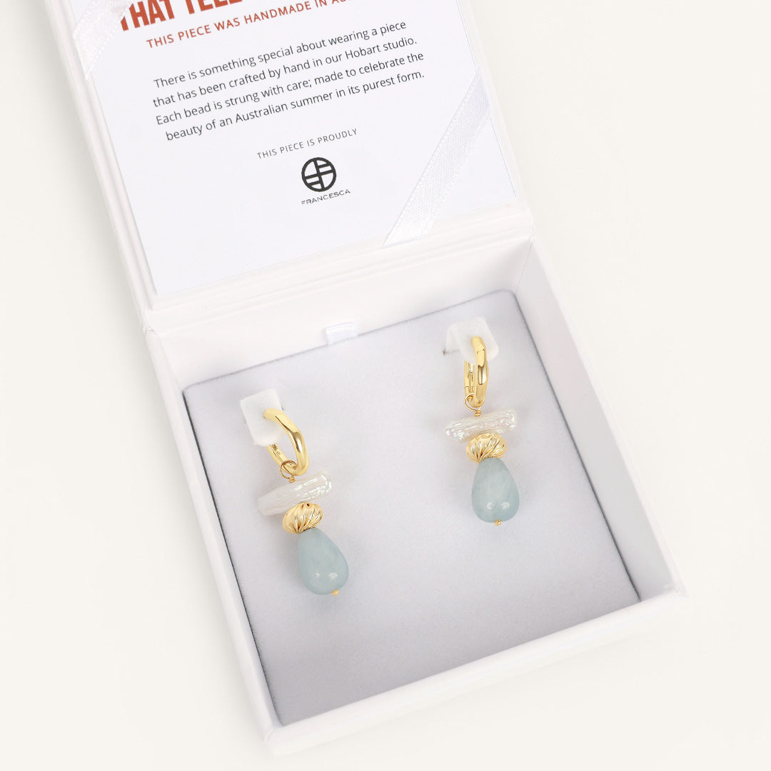 Pair of gold and light blue earrings in a box with a card on a white background