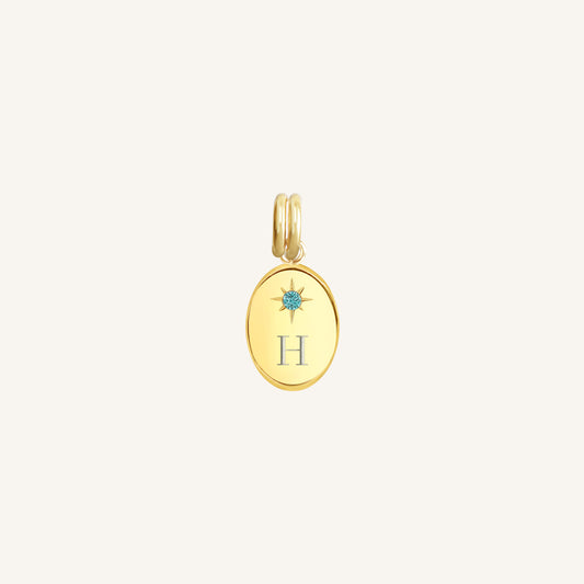 Gold pendant with 'H' and blue gemstone on a light beige background