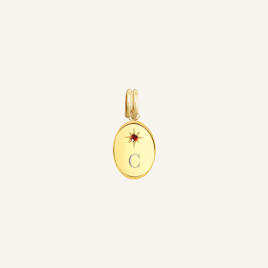 Gold pendant with letter 'C' and red stone on a light beige background